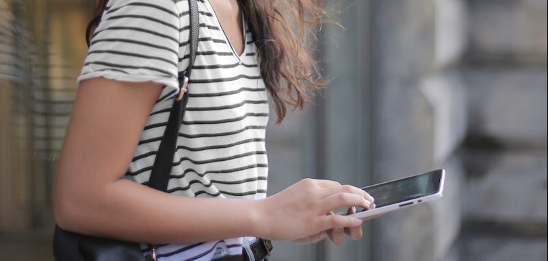 Side view of teen female in casual outfit and glasses browsing smartphone while smiling standing on street