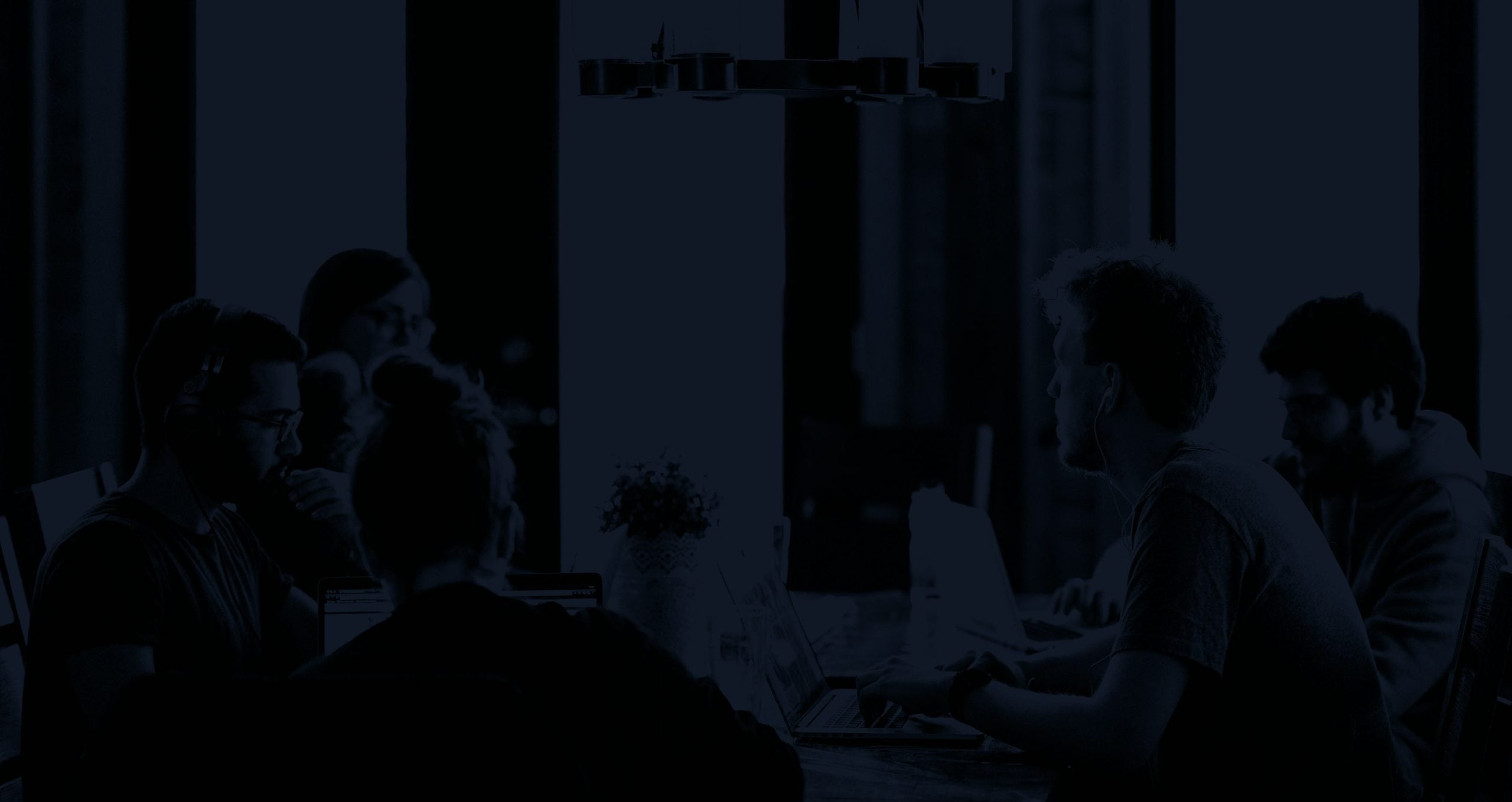 A dark, blue-toned image of several people sitting around a table with laptops in a dimly lit room.