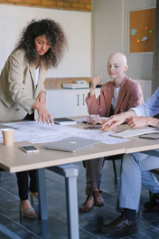 A group of colleagues discusses sketches and notes pinned to a mobile whiteboard in an office.