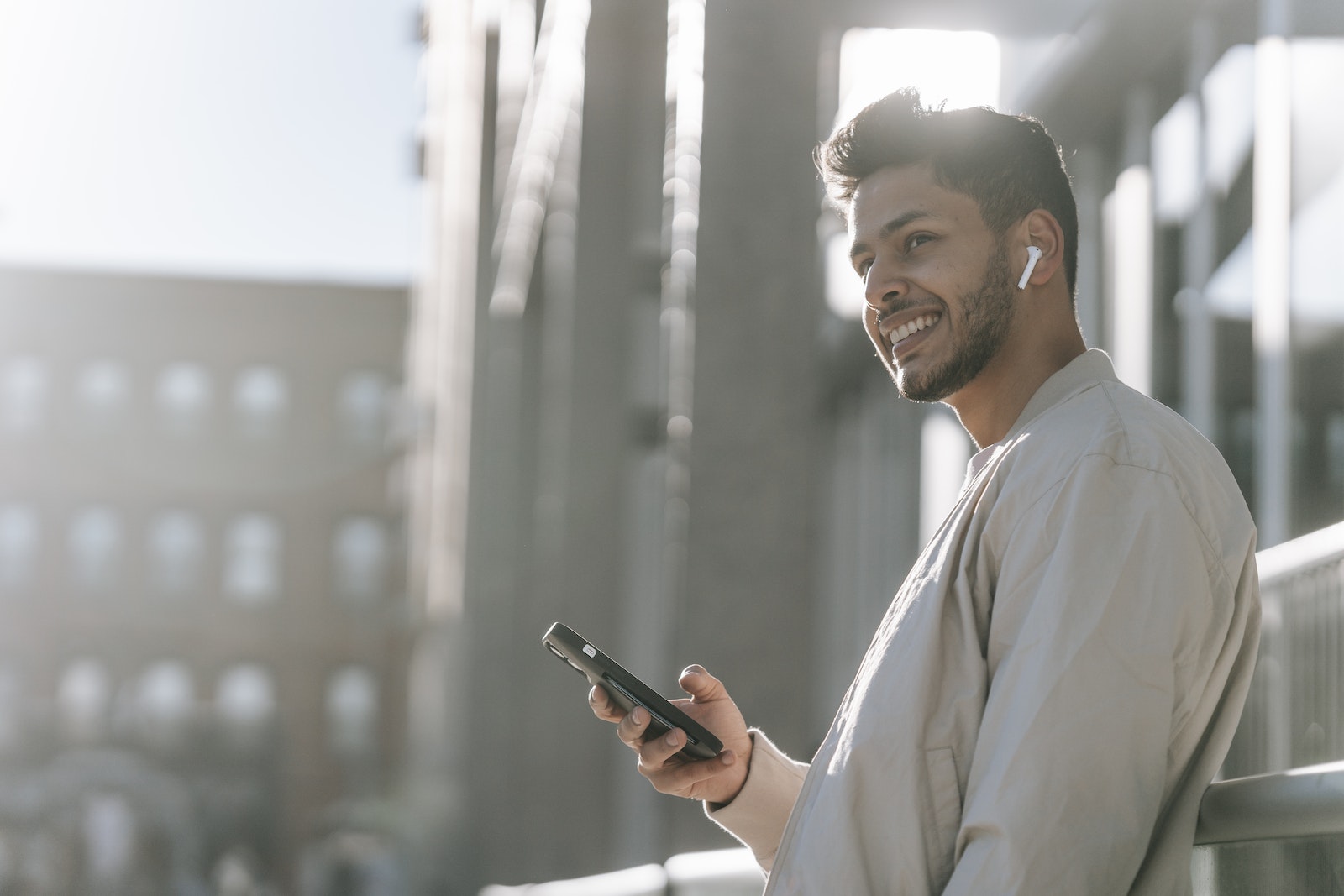 Side view of cheerful young bearded Hispanic male in TWS earbuds using mobile phone while standing near modern urban building in sunny day
