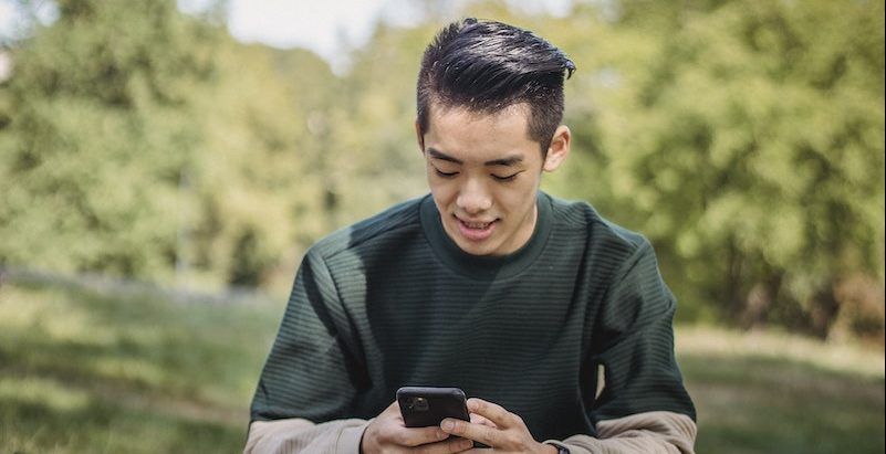 Smiling young ethnic man searching information in cellphone while sitting on stone in park with takeaway pizza