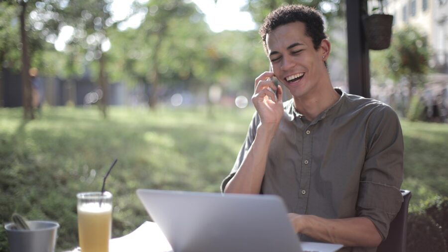 Content African American male freelancer wearing casual clothes sitting at table in street cafe with laptop and speaking on cellphone while working on startup