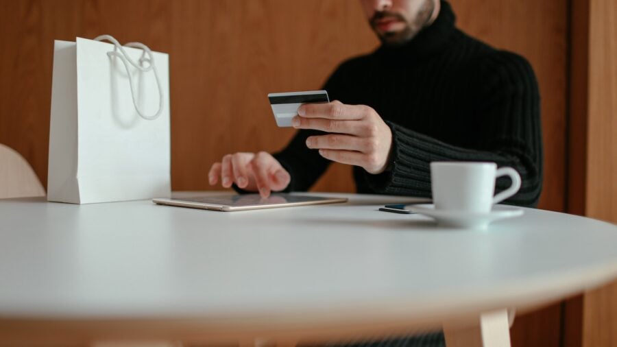 Crop focused bearded young man using tablet while doing online purchase with credit card sitting at table in modern cafe with coffee cup and shopping bag